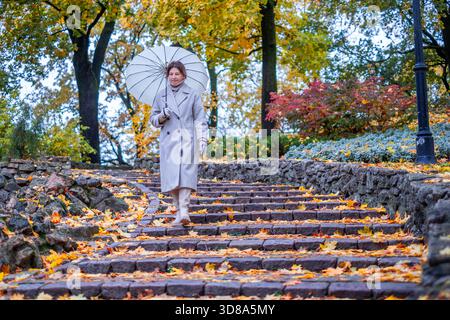 Woman in elegant coat walks down stone steps in a vibrant autumn park, holding a white umbrella, surrounded by fallen leaves and natural beauty Stock Photo