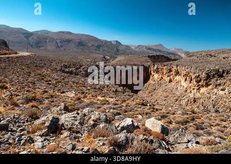 Wide rocky gorge near Xerokampos on Crete with rugged orange cliffs, dry shrubs and mountain slopes under clear blue sky. Harsh Mediterranean landscap Stock Photo