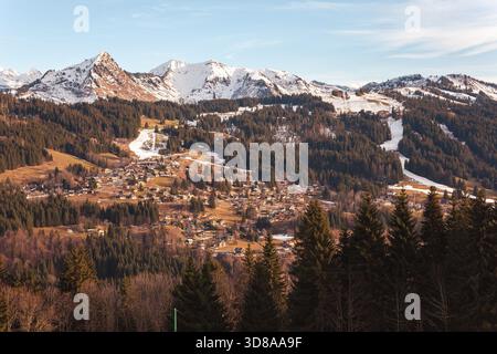 Les Gets village sits among forested hills and snowy alpine peaks in the French Alps on a clear winter day, showing chalets, ski slopes and mountains. Stock Photo