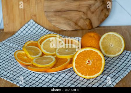 Sliced oranges and lemons on white plate with halved citrus fruits on checkered napkin and wooden cutting boards in kitchen. Concept of healthy food, Stock Photo