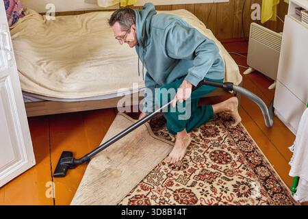 Man standing on one knee using vacuum cleaner to clean floor under lifted carpet in bedroom, smiling slightly. Stock Photo
