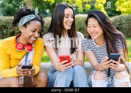 Three young women laughing and using smartphones outdoors in summer Stock Photo