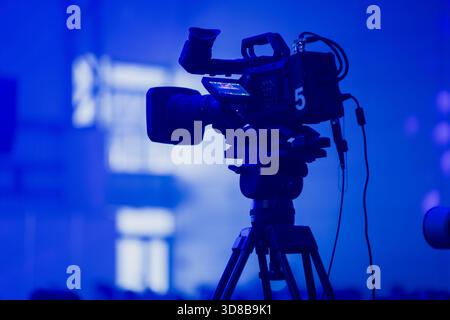 Professional broadcast camera mounted on a tripod and illuminated by deep blue event lighting, highlighting technical equipment used in modern live pr Stock Photo