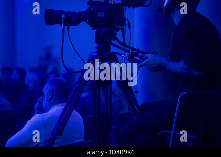Camera operator filming an event from a tripod platform under deep blue lighting with the audience in the background, illustrating professional event Stock Photo