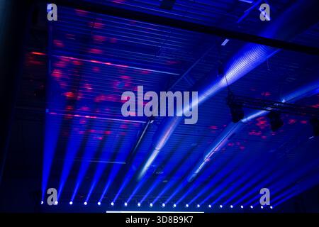 Strong blue stage wash beams projecting toward a metal ceiling with visible HVAC piping and truss rigging inside a modern live event production hall Stock Photo