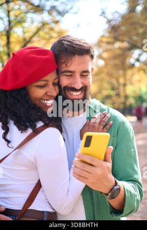 Interracial couple hugging and smiling while looking at smartphone in autumn park Stock Photo