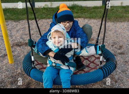 Two brothers in winter jackets sitting together on round nest swing at playground, older boy hugging younger sibling on cold autumn day. Concept of Stock Photo