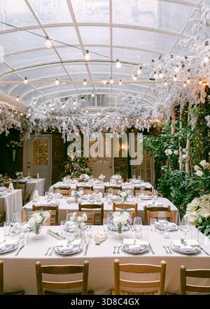 Tables are elegantly set in a glass greenhouse, adorned with winter decor and greenery for a wedding. Stock Photo