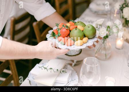 Fresh strawberries, green apples, and citrus fruits are placed on a lace-covered plate at a stylish table setting. Stock Photo