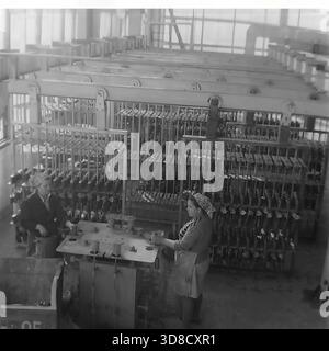 Slavyansk, Ukraine, USSR - 1980s: Interior view of the galvanic workshop at the AIZ Armature-Insulator Plant. Two female workers in aprons and headscarves stand at a loading station, manually attaching metal fittings to suspension racks for the electroplating process. Behind them, a massive automated overhead conveyor system moves thousands of parts through the line. This black and white archival photo captures the combination of manual labor and industrial automation in the peaceful Donbas city. Stock Photo