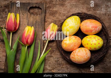 Festively decorated Easter eggs in vibrant colors beside blooming tulips on a rustic wooden table, capturing the essence of the Easter holiday spirit. Stock Photo