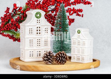 Two ceramic Christmas village houses with pine cones, red holly berries, and small tree on wooden tray on white textured background. Winter holiday ho Stock Photo