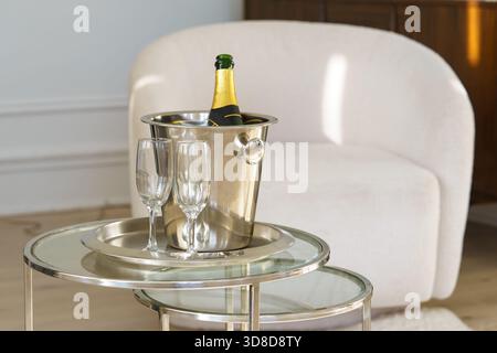 Champagne bottle in a metal ice bucket with two tall glasses on stylish glass table in cozy white interior background. Holidays, festive evenings, rom Stock Photo