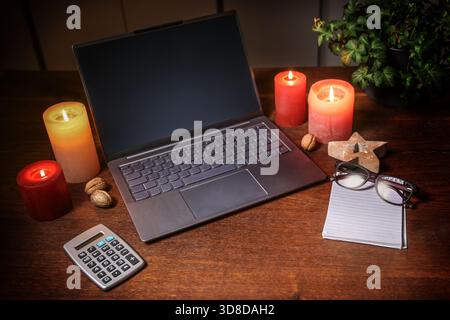 Laptop with empty screen, calculator and a notepad with glasses between candles and Christmas decoration on a dark brown wooden desk, online shopping, Stock Photo