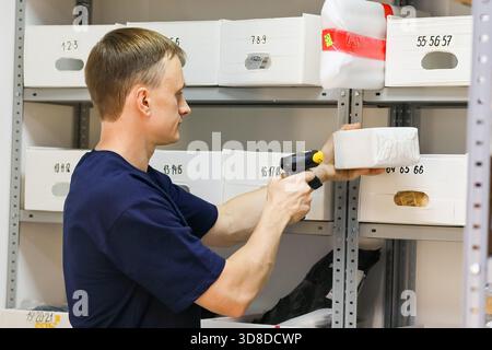 Side profile of man using handheld scanner on small white parcel beside labeled storage boxes. Stock Photo