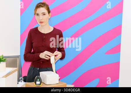 Worker prepares to seal white parcel with adhesive tape on cardboard box beside barcode scanner against colorful wall Stock Photo