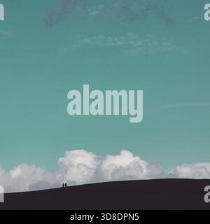 two small figures walking up a hill silhouetted against large storm clouds but with a clear green blue sky Stock Photo