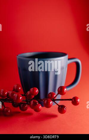 A deep blue mug stands against a bold red backdrop as glossy red berries hang above it, creating a clean, modern, and festive visual mood. Stock Photo