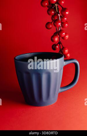 A deep blue mug stands against a bold red backdrop as glossy red berries hang above it, creating a clean, modern, and festive visual mood. Stock Photo