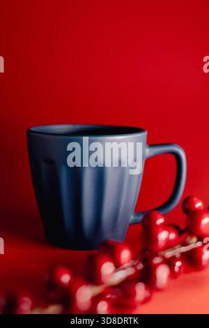A deep blue mug stands against a bold red backdrop as glossy red berries hang above it, creating a clean, modern, and festive visual mood. Stock Photo