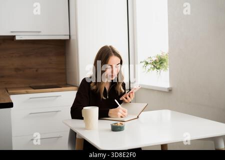 Young woman using smartphone writing notes in the notebook. doing planning and assignment. Stock Photo
