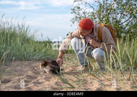 Caring man stroke petting cat on beach helping overcome fear of unfamiliar place. Cautious pet trust to owner, pet owner reassuring feline, human animal bond, nervous scared animal in new surroundings || Model released Stock Photo