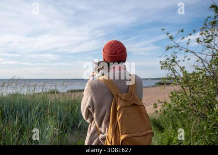 Rearview of grumpy frowning sullen cat presses close to male owner during outdoor walk. Reluctant affection, feline forced comfort seeking, animal bonding, pet owner reassurance in protective embrace. || Model released Stock Photo