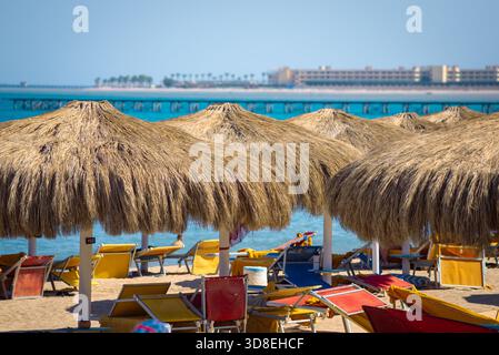 Rows of large straw beach umbrellas cast deep shade over colorful red, yellow, and blue sun loungers on sandy Hurghada shore, with turquoise sea, dist Stock Photo