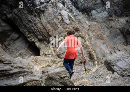 Young person climbing across rugged coastal rock formations, wearing a bright orange shirt, exploring a rocky shoreline outdoors. Stock Photo