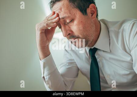 Troubled, exhausted businessman with a hand on his forehead, feeling overwhelmed by stress and fatigue. Selective focus. Stock Photo