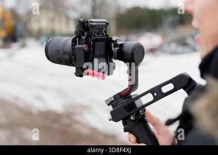 Videographer operating a mirrorless camera mounted on a gimbal stabilizer to capture stable footage in cold outdoor working conditions Stock Photo