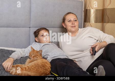 Mother and son relaxing on a bed watching TV with their sleeping ginger cat. for family, leisure, domestic lifestyle, and pet themes. Stock Photo