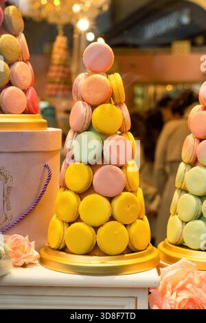 A vibrant cone of pastel macarons stacks into a confectionery tower at a shop display, evoking sweet treats and holiday joy. Warm lighting highlights Stock Photo