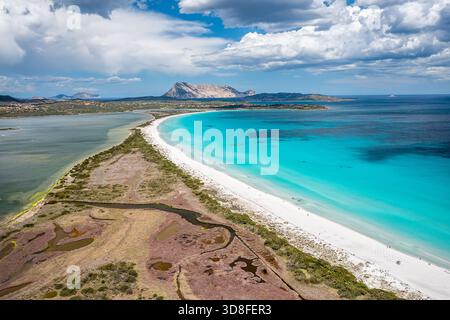 A pristine, curved beach with turquoise water and white sand meets calm seas along a coastal landscape under a blue sky. Distant island and mountains Stock Photo