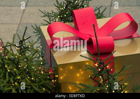 A large golden gift box with a red ribbon sits among small decorated Christmas trees with glowing fairy lights Stock Photo