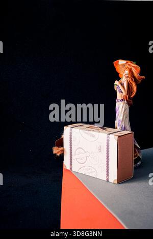 Decorative suitcase-style gift box with female figurine on orange and grey geometric surface, black background and copy space for travel concepts. Stock Photo