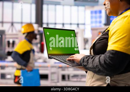 Female engineer holds laptop with mockup template, overseeing robotic arm automation in smart manufacturing facility. The industrial plant uses industry 4.0 software and conveyor belts for efficiency. Stock Photo