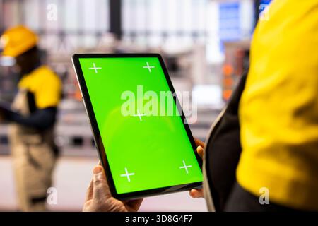 An engineer in industrial workspace holds tablet with a green screen, highlighting innovation and sustainability. The isolated chromakey display offers a blank template for industry use. Stock Photo