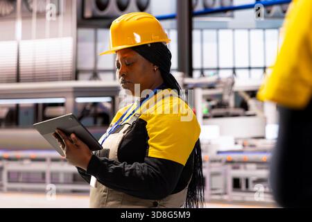 Professional woman wearing industrial helmet, managing high tech equipment through tablet in industry 4.0 plant. Quality assurance engineer ensuring smooth operations, efficiency and innovation. Stock Photo