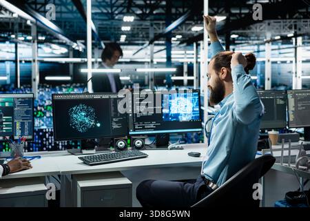 Data center system administrator stretching at PC desk to relieve back fatigue while using AI assistant to perform maintenance. Server room employee relaxing sore muscles after long hours at computer Stock Photo
