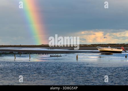 A small boat rests on the shore with a colorful rainbow stretching across a dramatic sky. Stock Photo