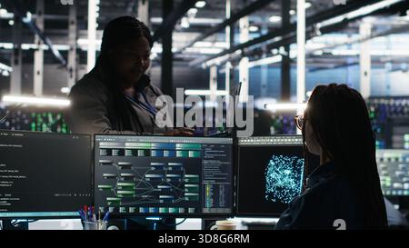 Data center manager supervising technician using node tree software to analyze datasets. Server room team leader oversees IT expert using visual programming tools to improve gear performance, camera B Stock Photo