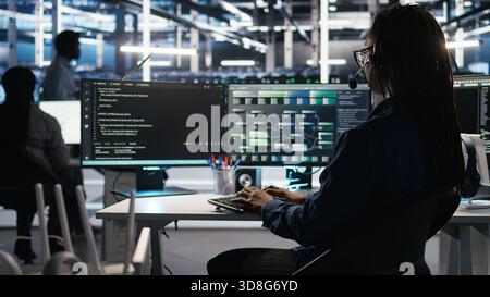 Data center employee uses audio headset and visual programming dashboard to troubleshoot client reported issues. Server farm technician looks at node tree interface, talking in microphone Stock Photo