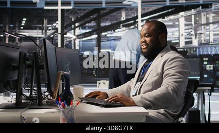 Joyous data center software developer delighted after solving all computer bugs. Smiling african american man in server hub pleased after finishing tasks on PC, overseeing server rigs, camera A Stock Photo