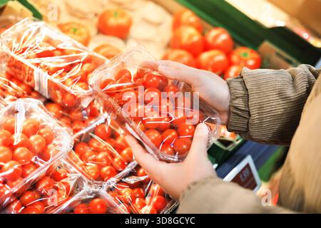 Close-up of a person's hands holding a clear plastic container of fresh cherry tomatoes in a grocery store. High quality photo Stock Photo