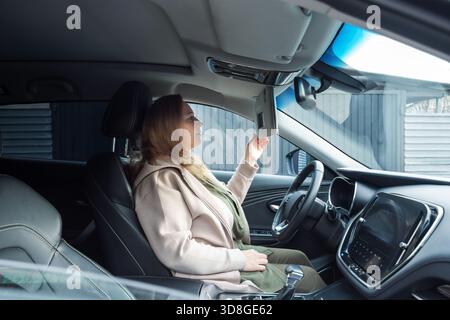 Adult woman checking the vanity mirror attached to the sun visor while sitting in the driver's seat of a modern car. for topics related to commuting, Stock Photo