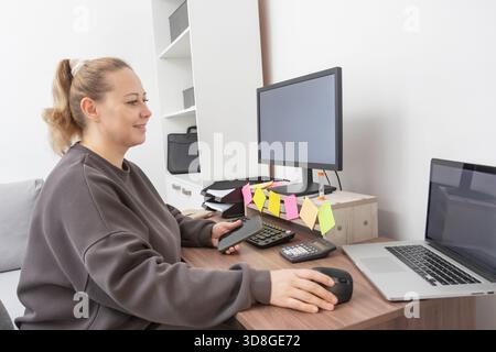 Caucasian middle-aged woman telecommuting from home office. She uses multiple screens and tools like a calculator and sticky notes. for illustrating p Stock Photo