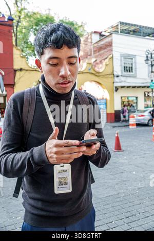 Mexico City Mexico,Coyoacan,young adult man Hispanic standing street using smartphone focused expression wearing black turtleneck backpack lanyard bad Stock Photo
