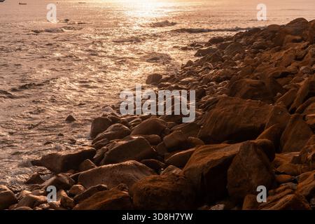 Warm, golden hour sunset reflecting on the ocean waves and the rough texture of large, brown, wet rocks on the coast. Stock Photo