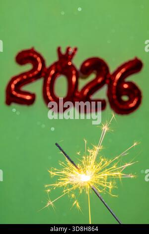 Festive New Year photo featuring a bright burning sparkler in the foreground with red foil “2026” balloons softly blurred in green background Stock Photo